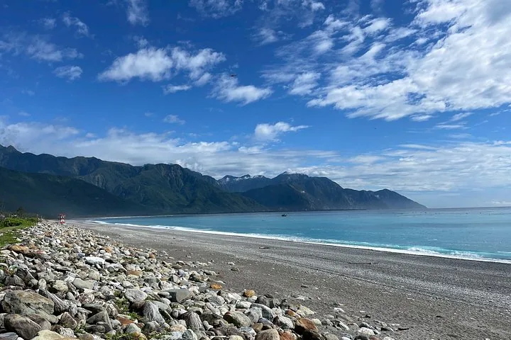 A wide rocky beach along the coast with lush green mountains in the distance.
